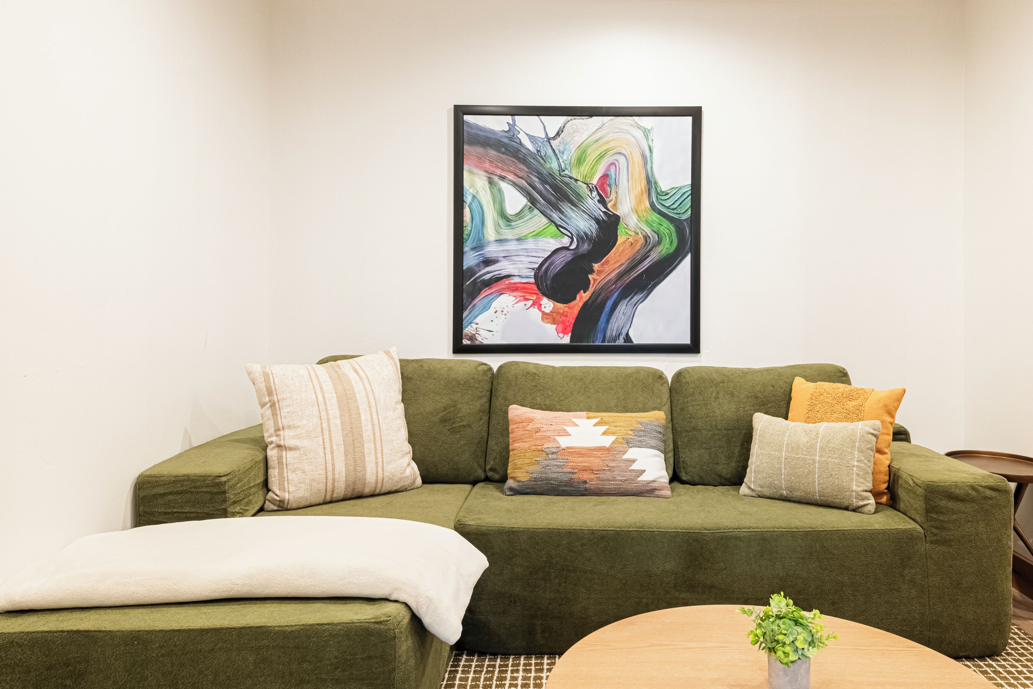 Un canapé vert agrémenté de plusieurs coussins, un tableau abstrait encadré et une petite plante posée sur une table en bois.