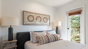 Un dormitorio ordenado con una cama grande, dos mesitas de noche con lámparas, un adorno en la pared y una puerta que da a un balcón con vegetación.