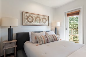 Um quarto bem arrumado com uma cama grande, duas mesas de cabeceira com abajures, um elemento decorativo na parede e uma porta que dá para uma varanda com vegetação.