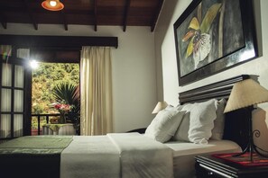 Een slaapkamer met een bed, nachtkastje, lamp, schilderij en uitzicht op een tuin door een raam.