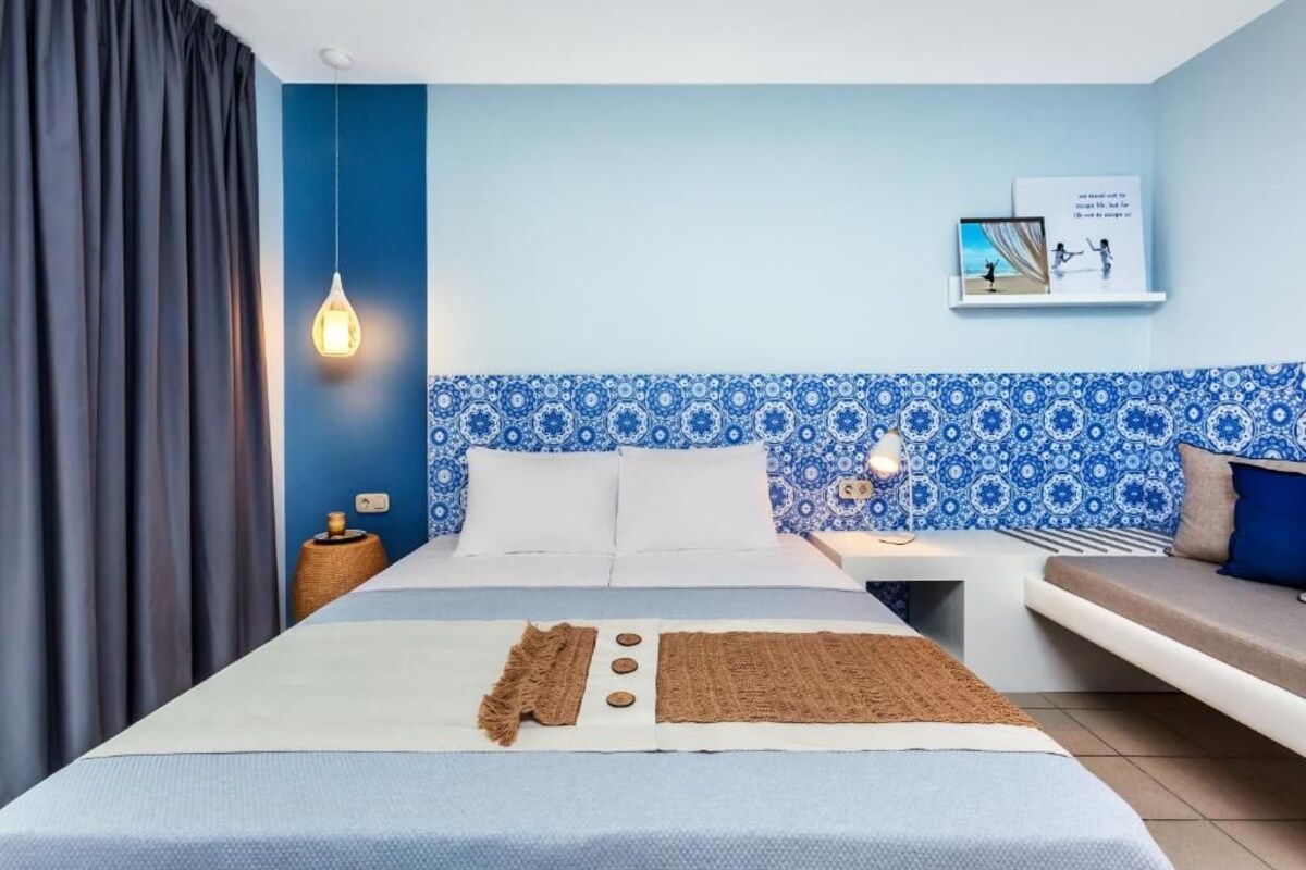 A modern hotel room with a large bed, a bedside table, a wall-mounted shelf with books, and a patterned blue and white tile backsplash.