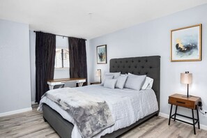 Quarto com cama, escrivaninha, cadeira, mesa de cabeceira e janela com cortinas.
