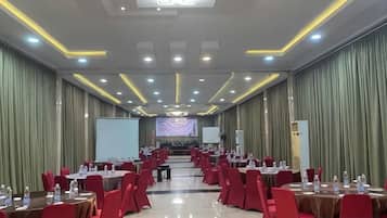 A conference room with rows of tables and chairs, a projector screen, and a stage at the front.