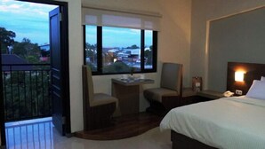 A hotel room with a bed, two chairs, a small table, a nightstand, and a window with a city view. - Bukit Indah Lestari Hotel (Tanjung Baru)