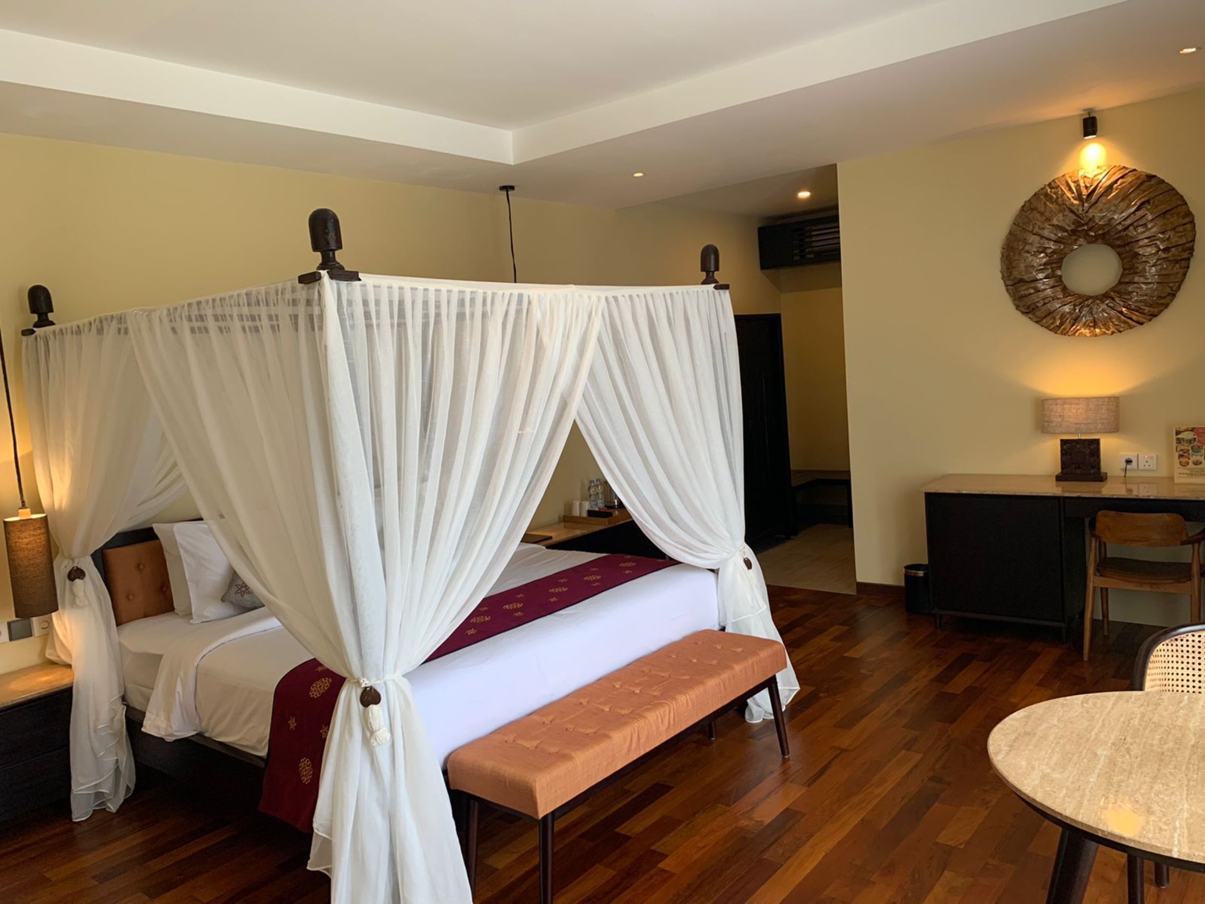 A four-poster canopy bed with a bench, a desk, and a round table in a hotel room.