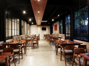 A spacious dining area with multiple tables and chairs, large windows with a view of greenery, and a modern ceiling design.