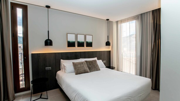 Ein modernes Hotelzimmer mit einem großen Bett, zwei Nachttischen, einer Wandleuchte und einem fenster mit Vorhängen.