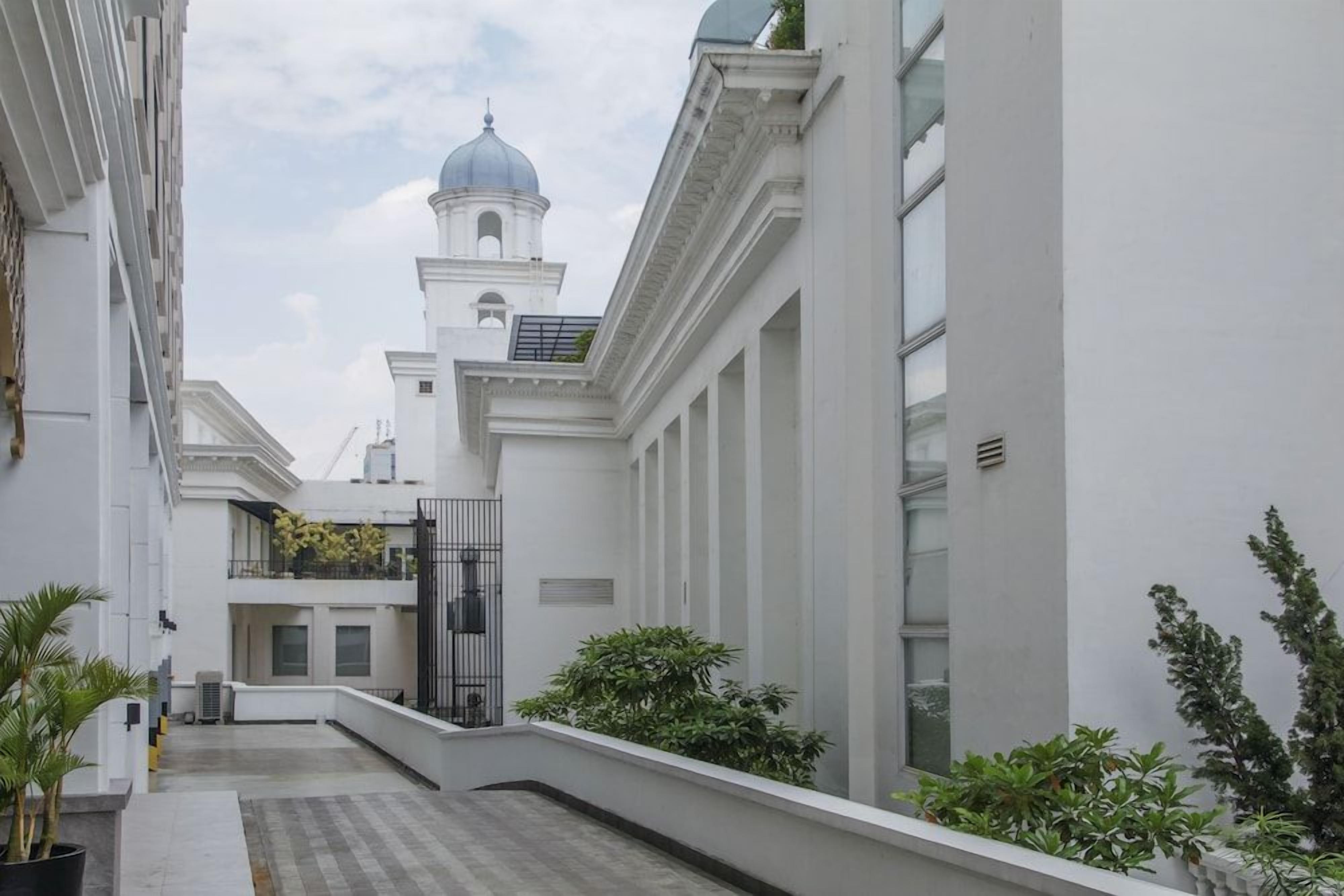 Une cour avec un bâtiment blanc, un petit jardin et le clocher d’une église qui se dresse au loin.