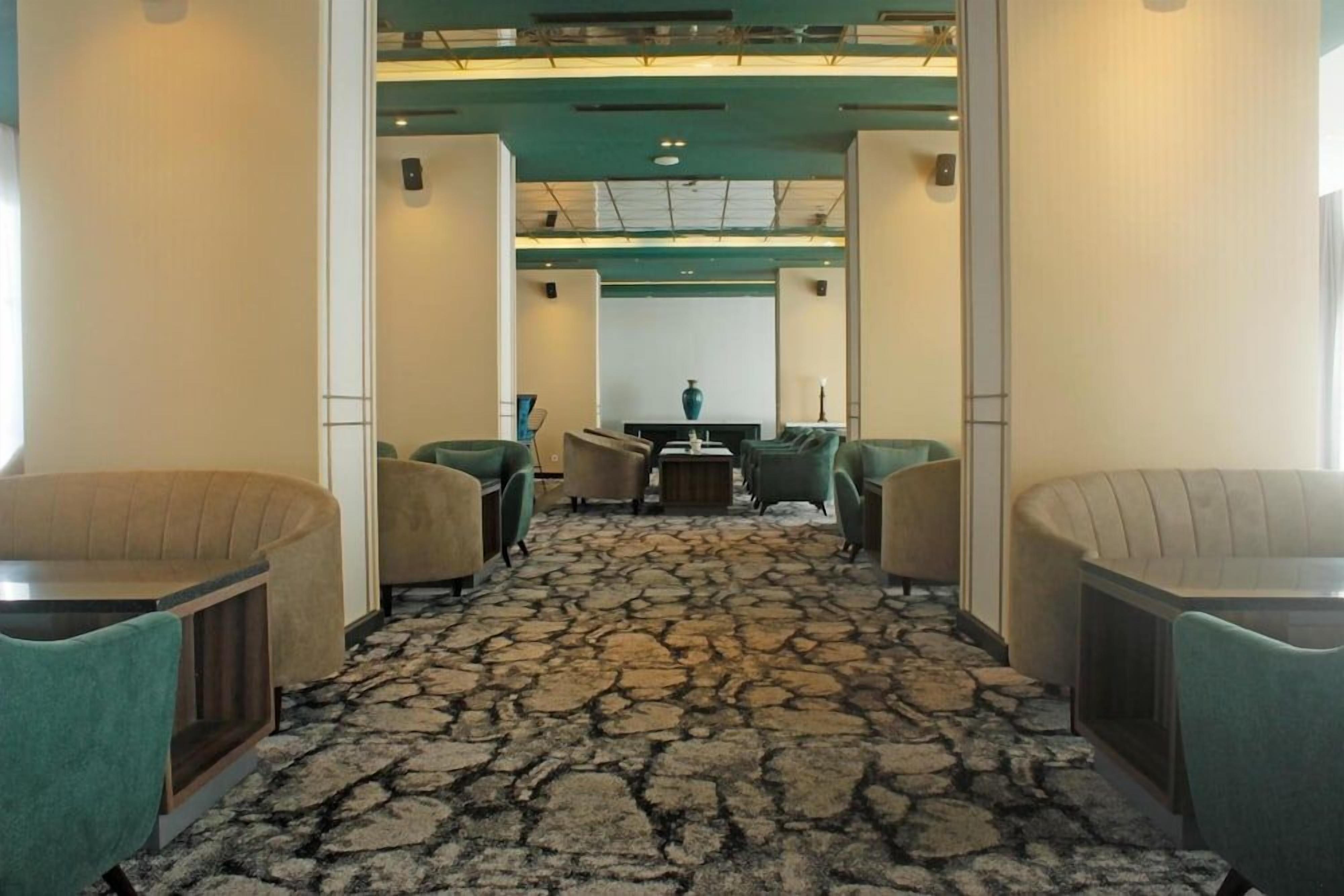 A hotel lobby with a central reception desk, green upholstered chairs, a patterned carpet, and a modern ceiling design.