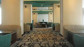 A hotel lobby with a central reception desk, green upholstered chairs, a patterned carpet, and a modern ceiling design.