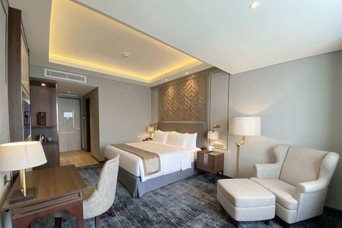 A hotel room with a bed, bedside tables, a desk with a chair, a sofa, and a carpeted floor.