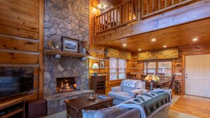 A cozy log cabin interior with a stone fireplace, wooden furniture, and a balcony.