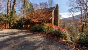 A rustic cabin with a stone chimney, surrounded by autumn foliage and a gravel driveway.