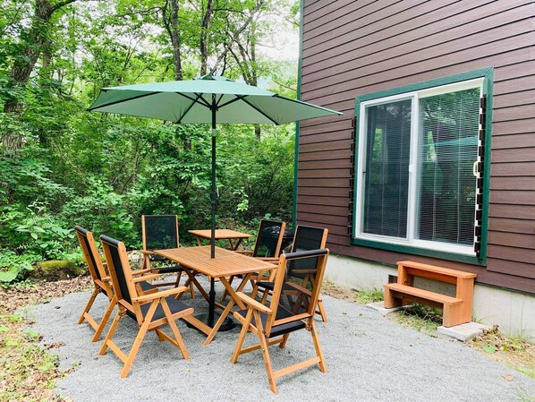 Un patio al aire libre con una mesa y sillas de madera, un paraguas verde y una ventana con persianas.