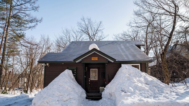 Una cabaña de color marrón oscuro, con puerta principal y un pequeño porche. Montones de nieve a ambos lados de la entrada.