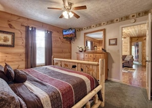 A wooden cabin bedroom with a bed, a ceiling fan, a television, and a dresser.