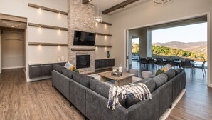 A modern living room with a large gray sectional sofa, a fireplace, a flat-screen TV, and a dining area with outdoor seating. - Luxury Retreat w/Views: Putting Green/Game Room/Infinity Pool & More! (Escondido)
