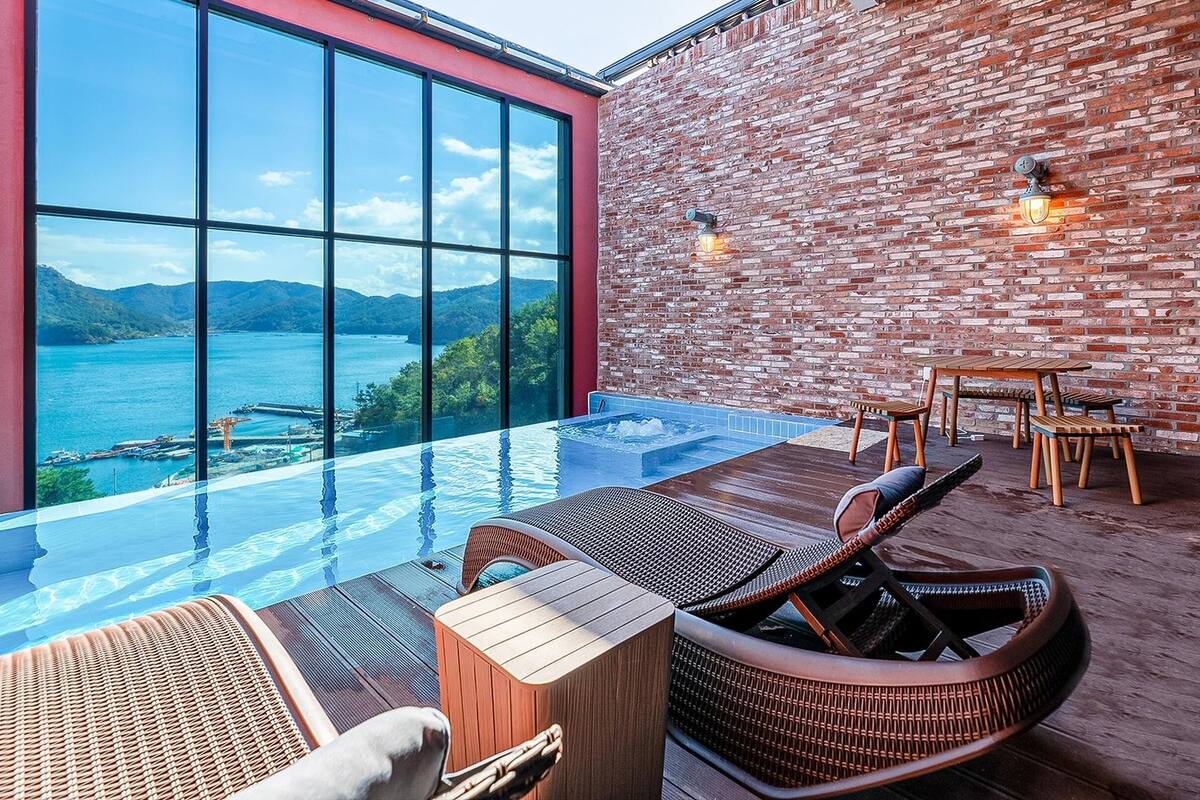 An infinity pool with a view of the sea and mountains, wooden furniture, and a brick wall.