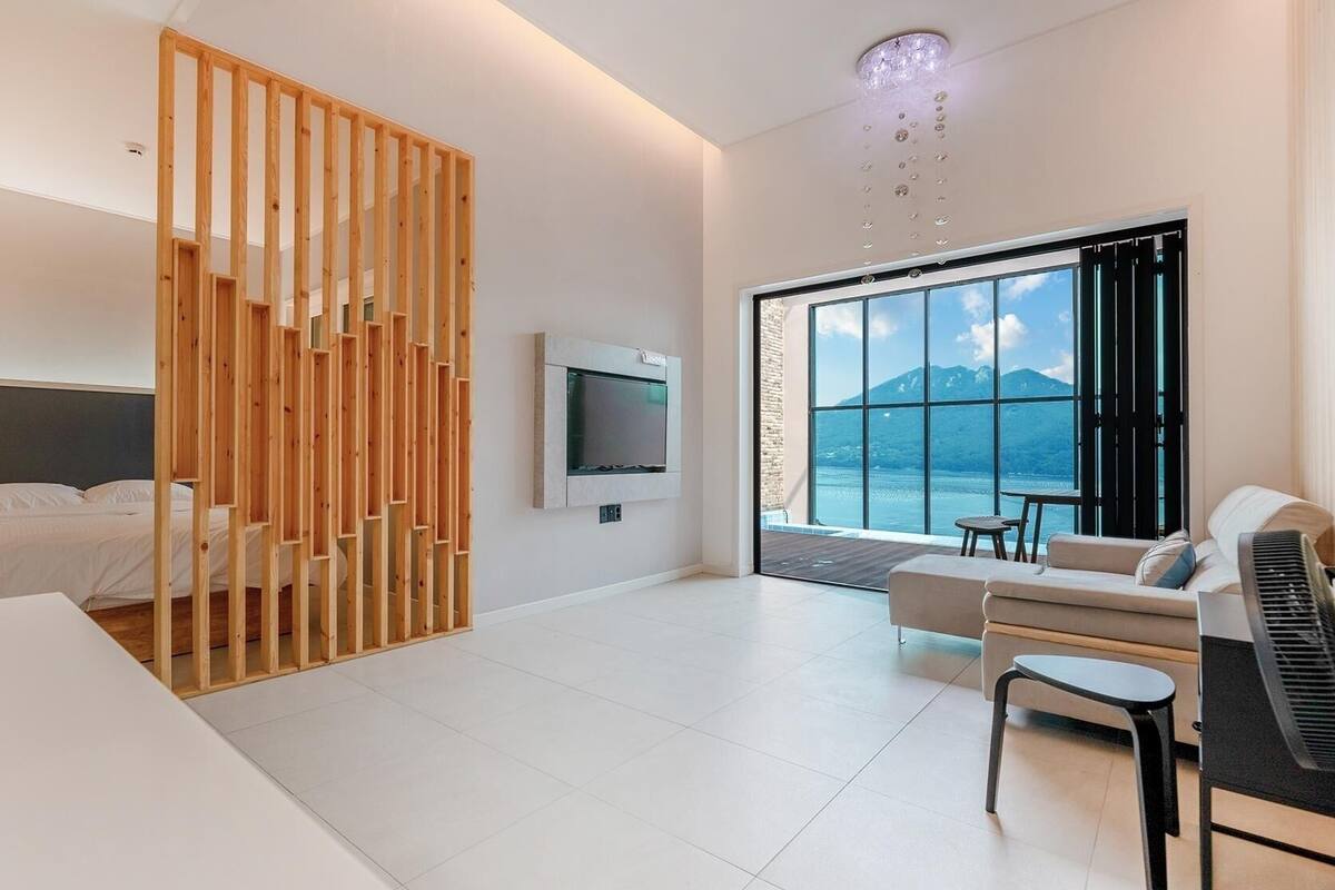 A modern hotel room with a bed, a TV, a piano, a sofa, and a large window with a mountain view.