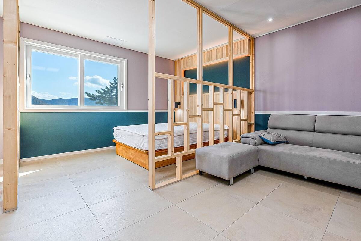 A bedroom with a bed, a grey sofa, a wooden partition, and a window with a view of mountains.