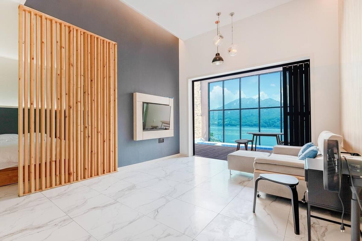 A modern hotel room with a bed, a TV, a sofa, a desk, and a large window with a view.