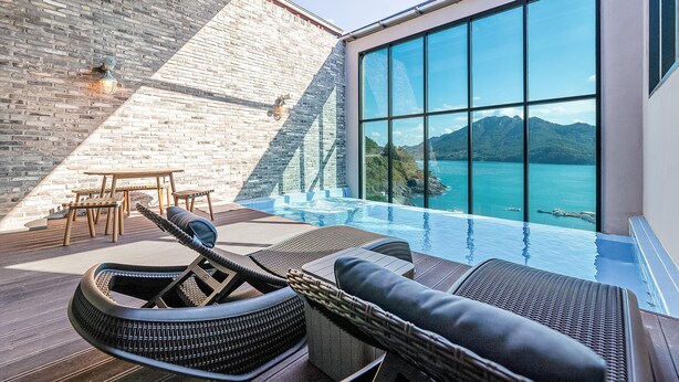 Una piscina infinita moderna con vistas a la montaña y al mar, muebles de madera y un muro de ladrillo.
