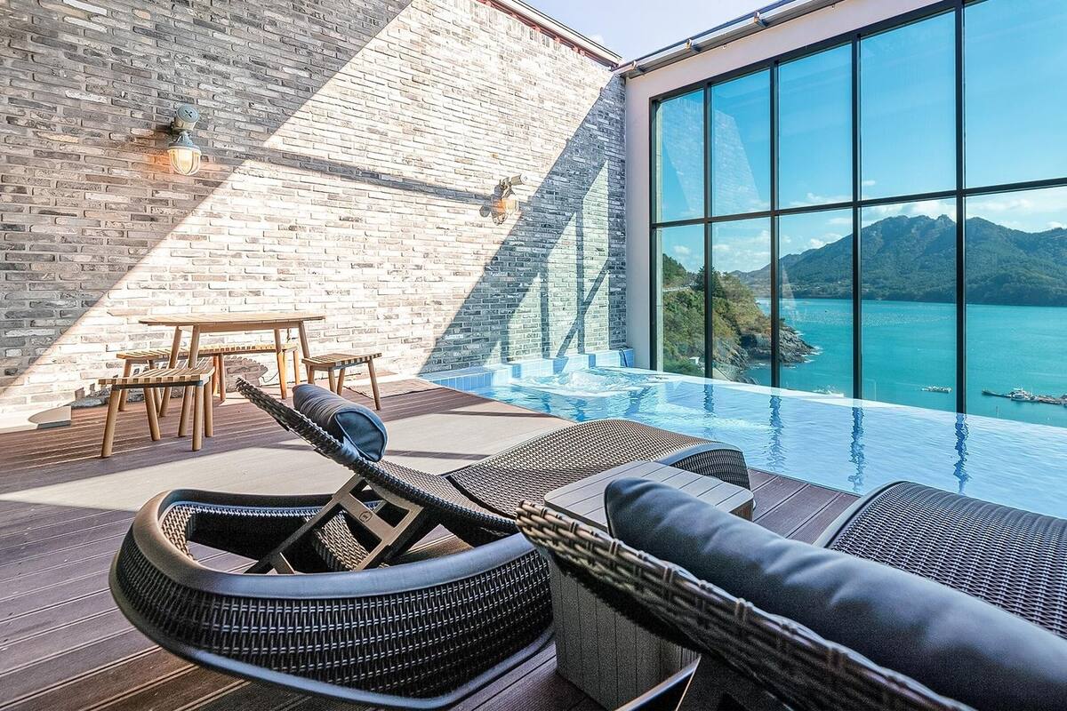 A modern infinity pool with a view of a mountain and sea, wooden furniture, and a brick wall.