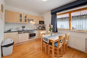 Eine kleine Küche mit Holzausbau, einem Esstisch mit Stühlen, einer Vase mit Blumen und einem Fenster mit Vorhängen.
