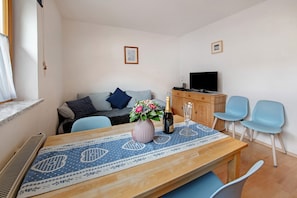 Ein Wohnzimmer mit einer Couch, einem Fernseher, einem Holztisch mit Tischdecke und zwei Stühlen.