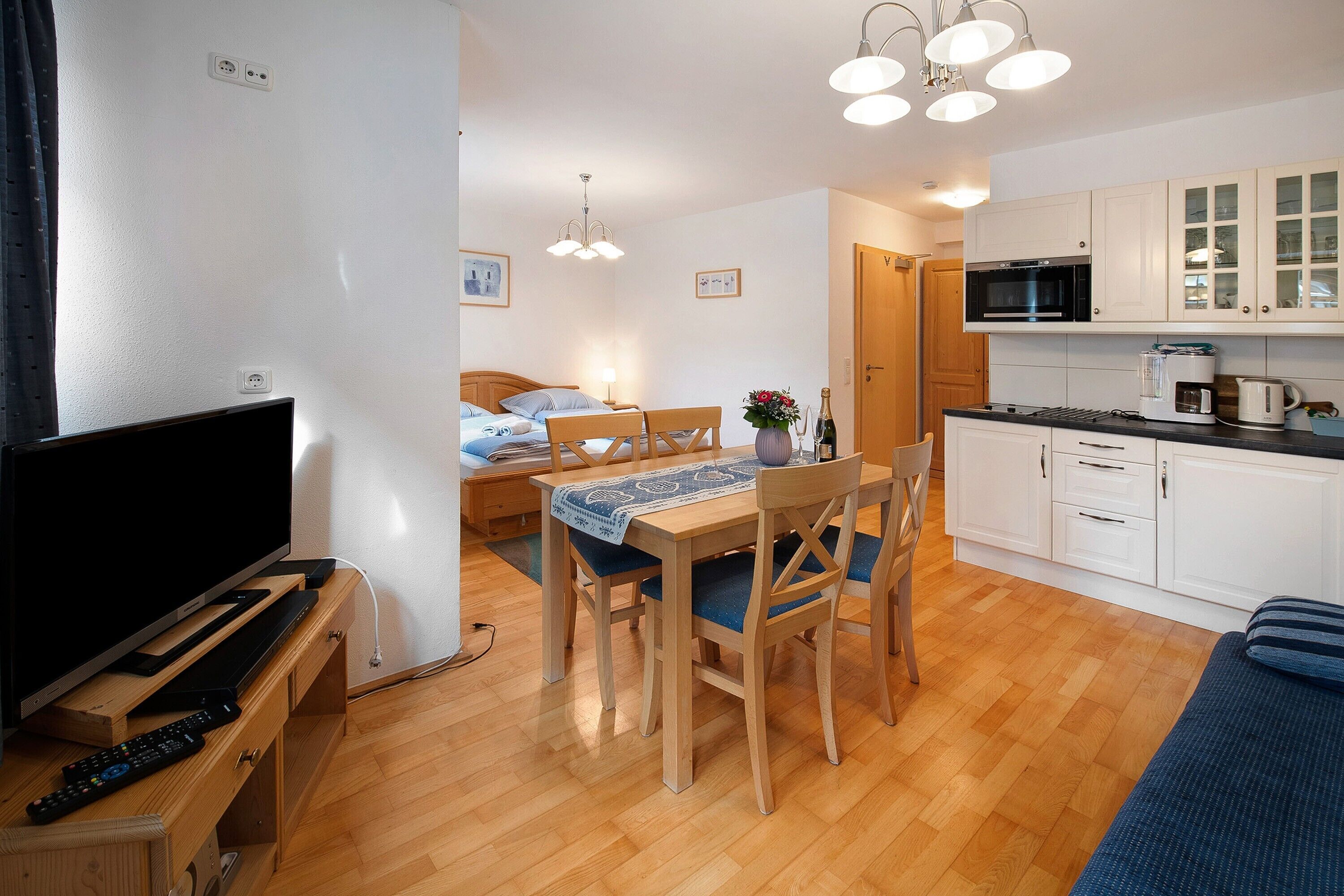 Une chambre d’hôtel compacte comprenant une télévision, une table à manger en bois, des chaises, un lit, une kitchenette et une porte donnant sur une autre pièce.