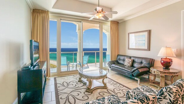 Habitación de hotel con balcón, sofá de cuero, mesa de centro de vidrio y televisor sobre un mueble.