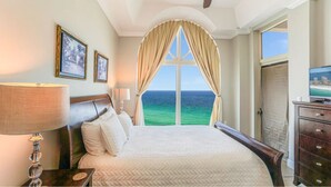 Habitación de hotel con cama, mesita de noche, lámpara, cómoda y un amplio ventanal con vista al océano.
