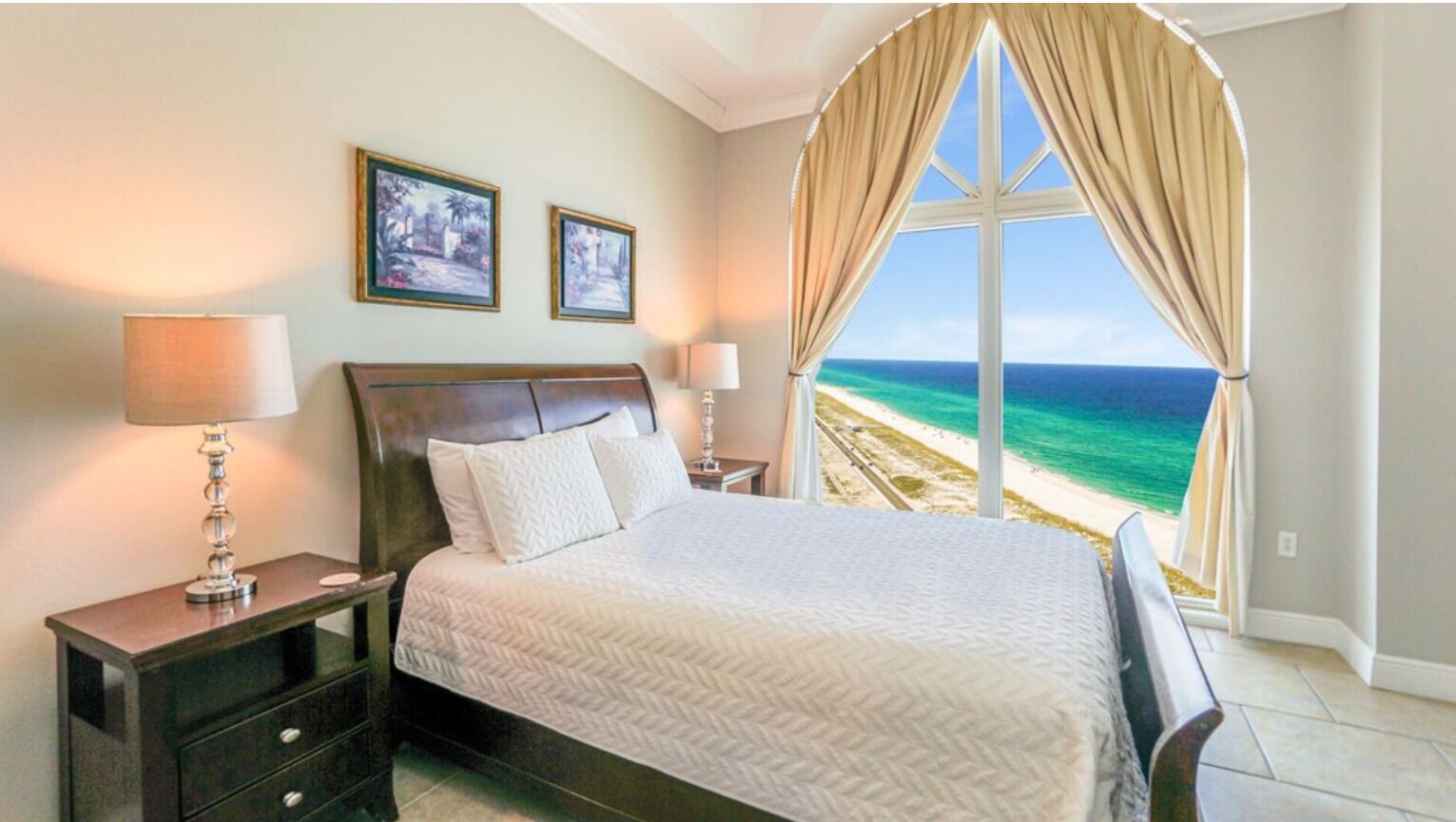 Une chambre avec un grand lit, deux lampes de chevet, une table de nuit et une vue sur la plage, visible à travers une grande fenêtre munie de rideaux.
