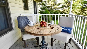 A wooden table with a glass bowl of apples and two white chairs with a blue pillow.