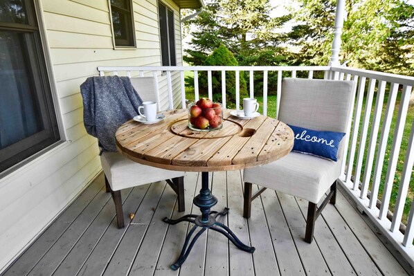 A wooden table with a glass bowl of apples and two white chairs with a blue pillow.