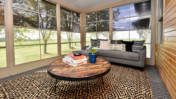 A room with large windows, a round wooden coffee table, a gray sofa with patterned cushions, and a blue potted plant.