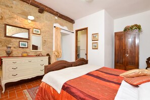 Une chambre avec un mur en pierre, une armoire en bois, un lit recouvert d’une courtepointe rouge et blanche, et une commode avec des tiroirs.