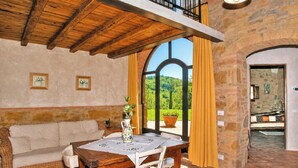 A cozy room with a wooden ceiling, a dining table with chairs, a sofa, and a view of greenery outside.