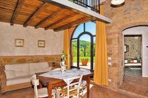 A cozy room with a wooden ceiling, a dining table with chairs, a sofa, and a view of greenery outside.
