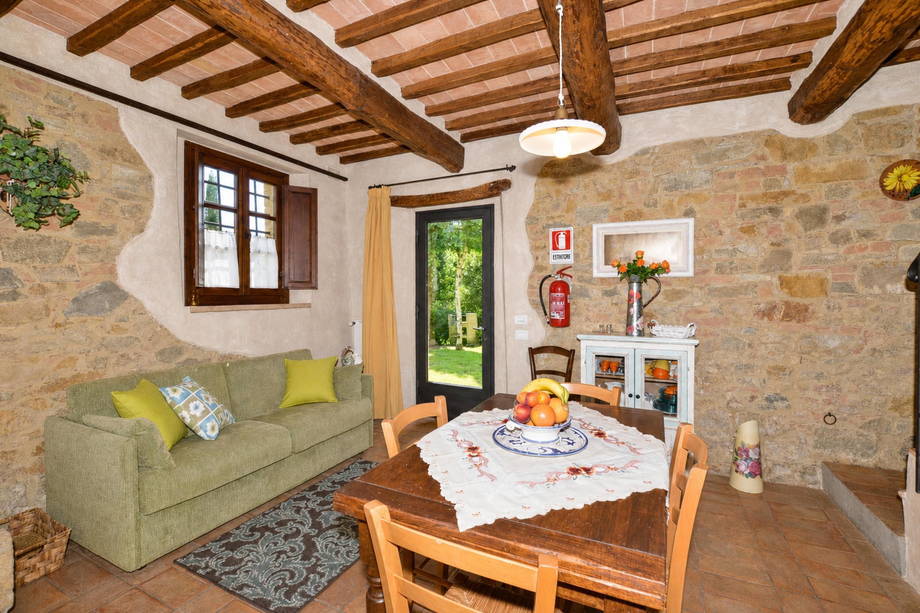 A rustic room with a stone wall, wooden beams, a green sofa, a dining table with a fruit centerpiece, and a fire extinguisher on the wall.