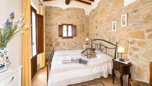 A bedroom with a stone wall, wooden beams, a bed with a nightstand, a window, and a vase with flowers.