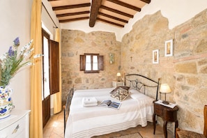 A bedroom with a stone wall, wooden beams, a bed with a nightstand, a window, and a vase with flowers.