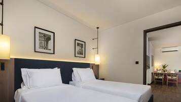A hotel room with two beds, a desk, and a chair. There are framed pictures on the wall and a lamp on the desk.