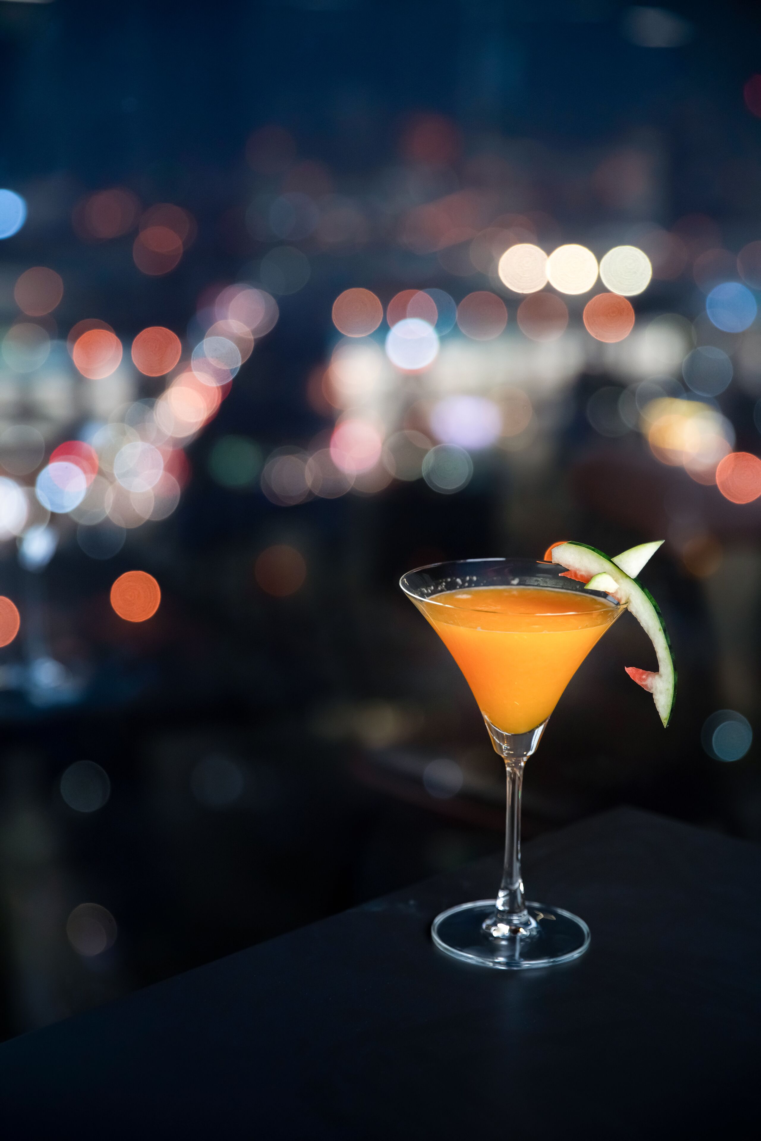 A cocktail in a martini glass with a garnish, set against a blurred cityscape background.