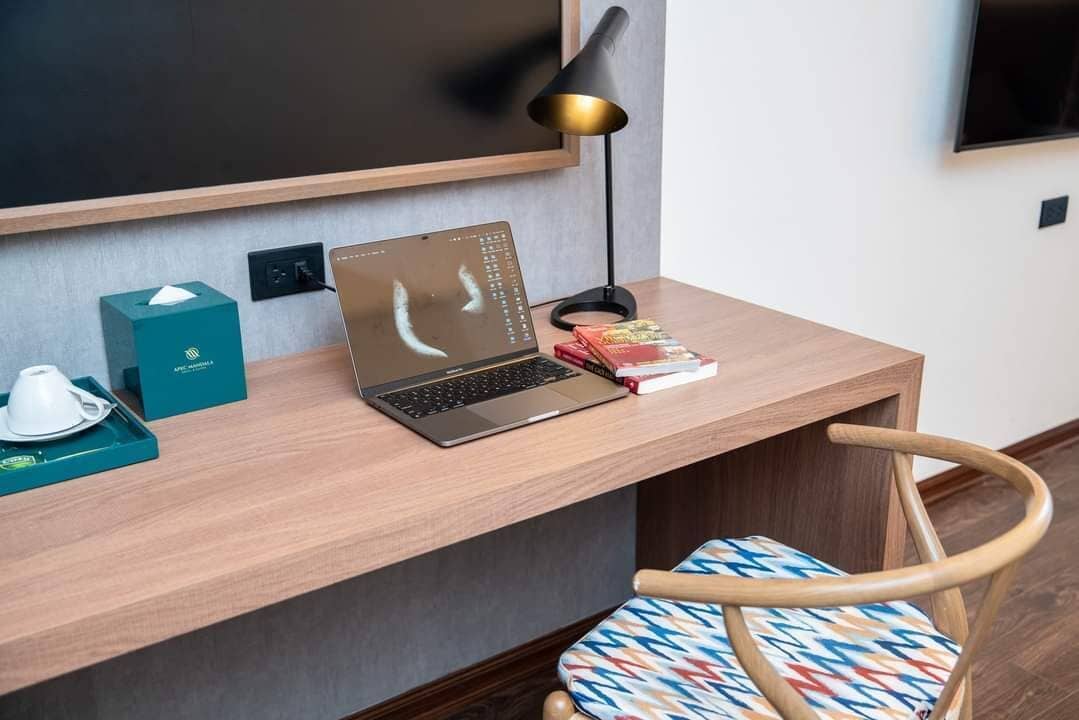 Un espacio de trabajo en un hotel con una computadora portátil, una caja de pañuelos, una bandeja con una taza, una lámpara y una silla.