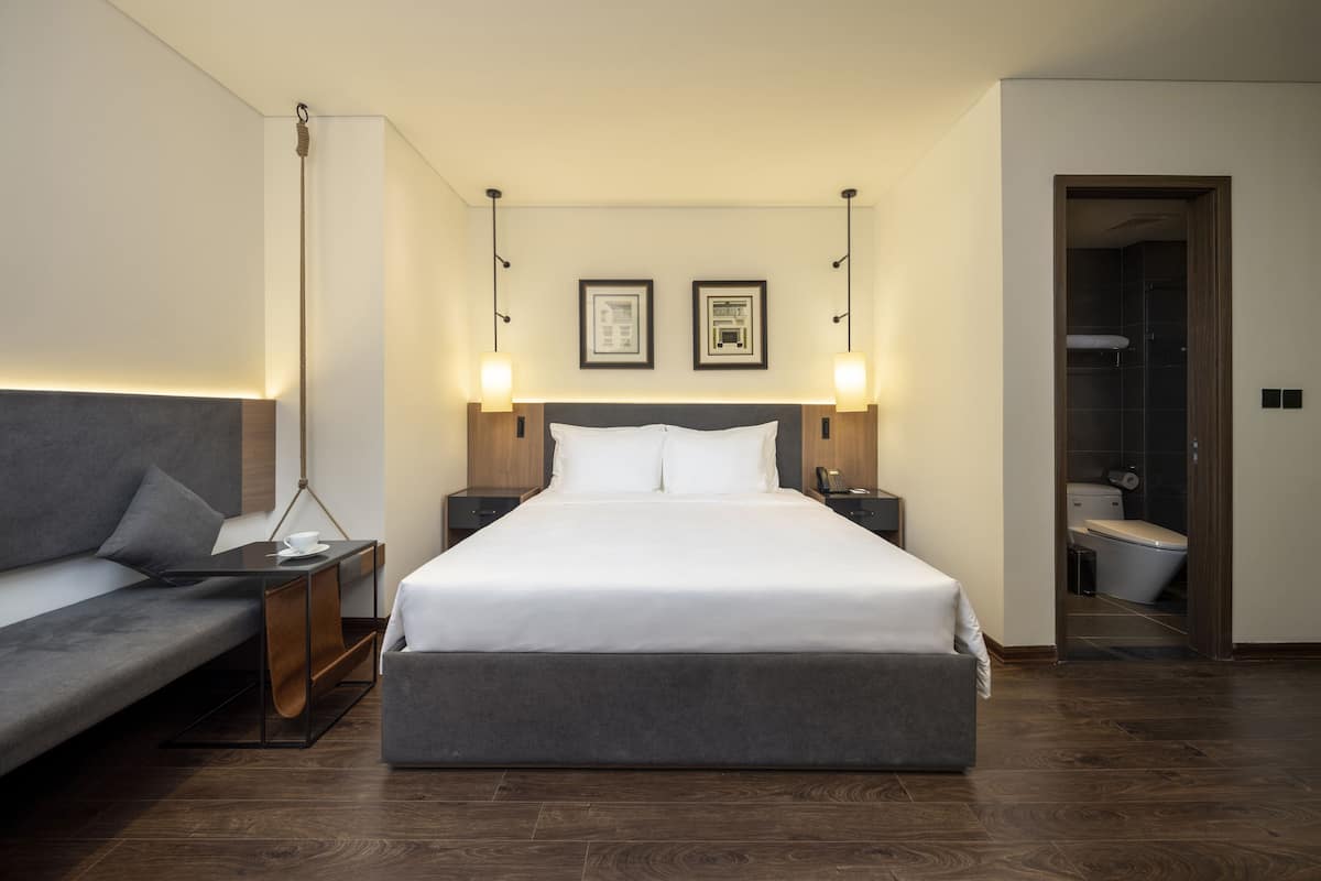 Una habitación de hotel moderna con una cama grande, un sofá, mesitas de noche y un baño visible a través de una puerta abierta.