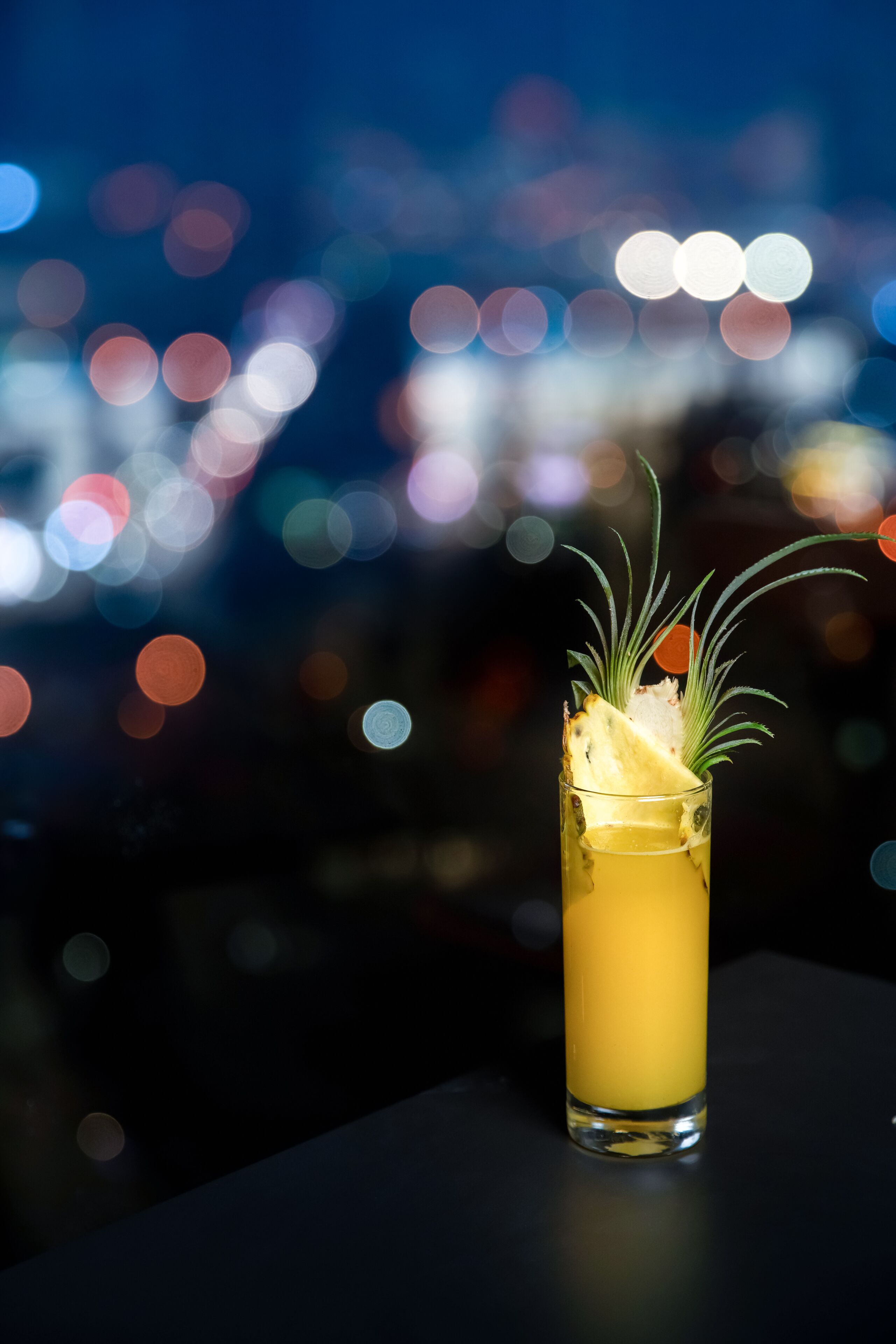 A tall glass with a yellow beverage, garnished with a slice of pineapple and a sprig of rosemary, set against a blurred cityscape background.