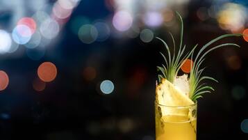A tall glass with a yellow beverage, garnished with a slice of pineapple and a sprig of rosemary, set against a blurred cityscape background.