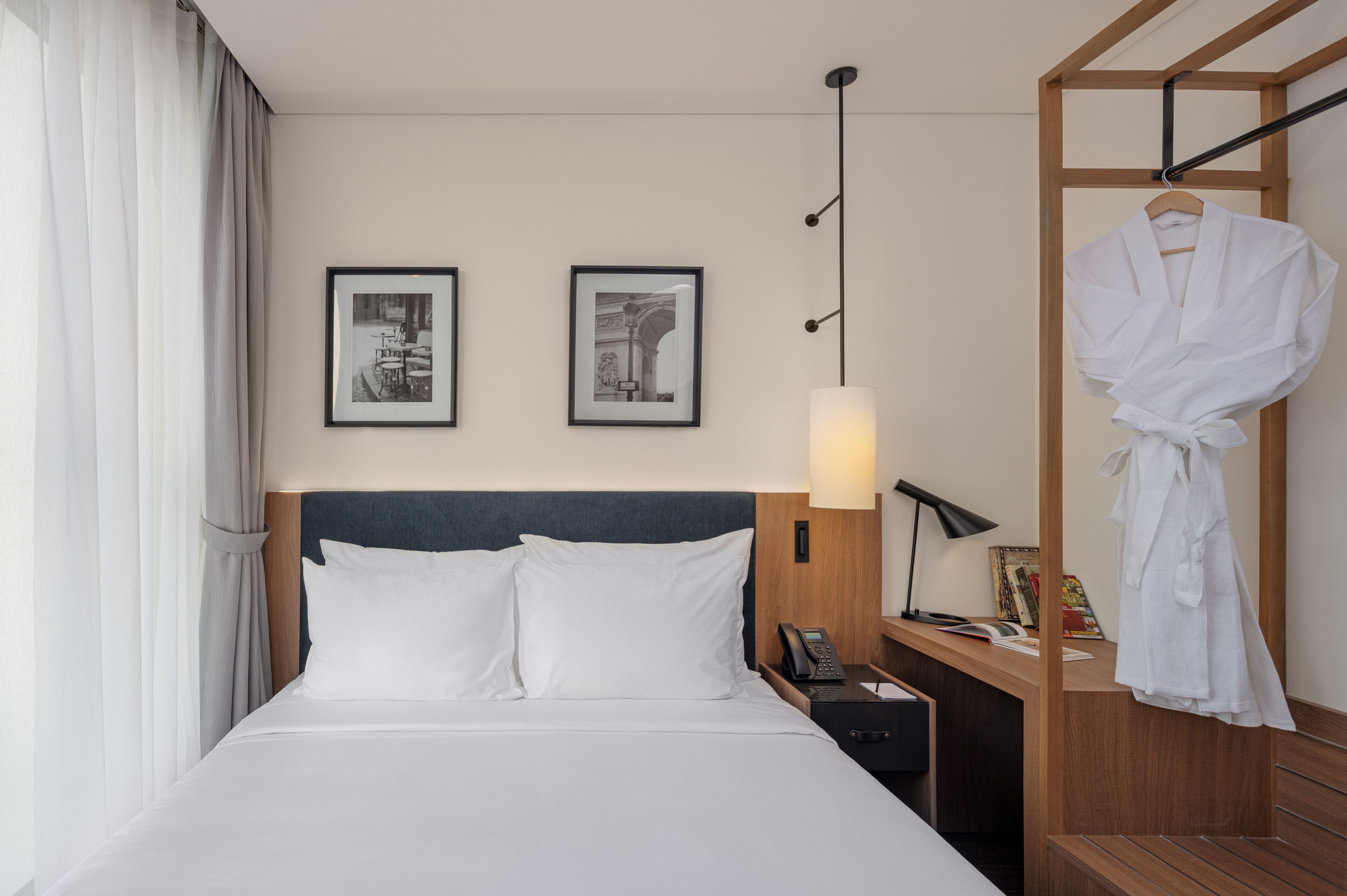 A hotel room with a bed, a nightstand with a lamp, a robe hanging on a rack, and two framed pictures on the wall.