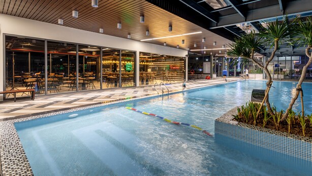 Una piscina interna con soffitto in legno, pareti di vetro e un’area relax con tavoli e sedie.
